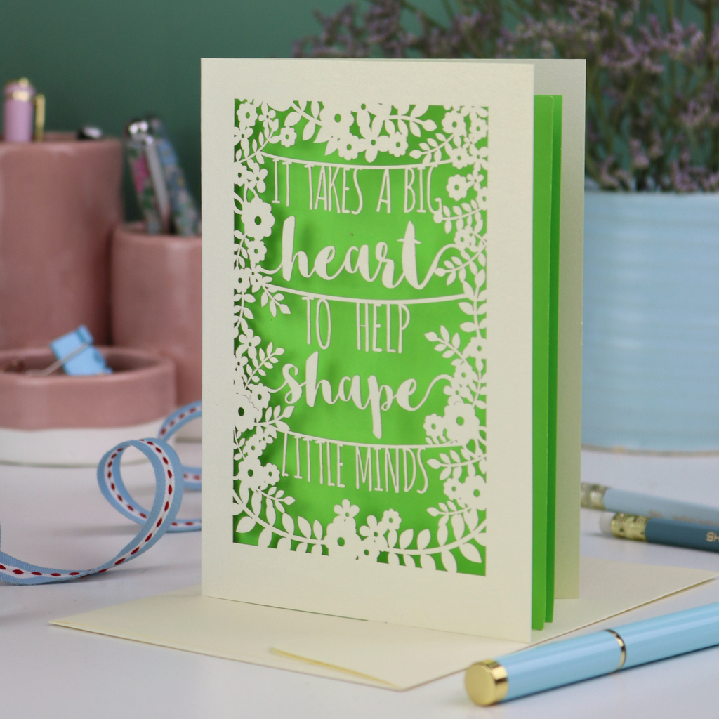A unique thank you teacher card with decorative text, 'It takes a big heart to help shape little minds' and floral patterns on a desk with stationery items.