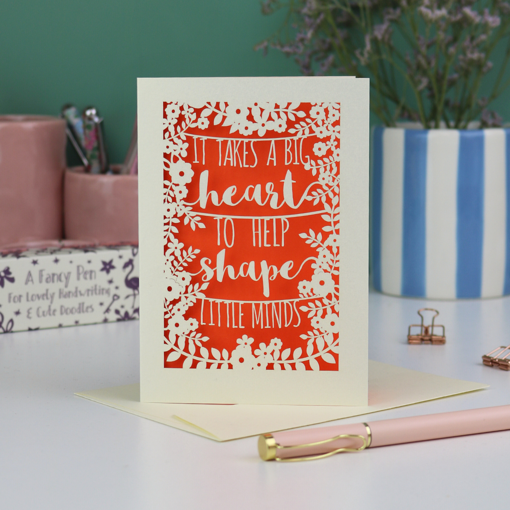 A handmade thank you teacher card with decorative text, 'It takes a big heart to help shape little minds' and floral patterns on a desk with stationery items.