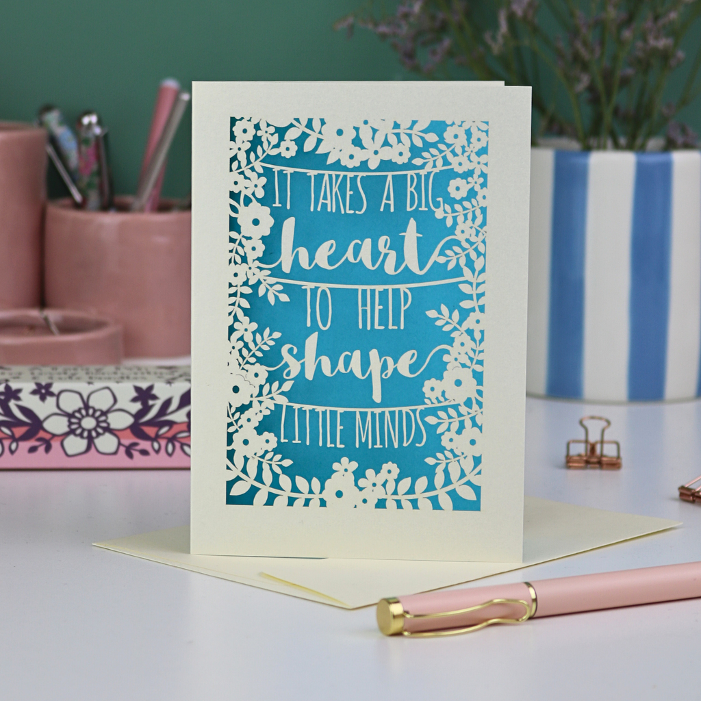 A keepsake thank you teacher card with decorative text, 'It takes a big heart to help shape little minds' and floral patterns on a desk with stationery items.