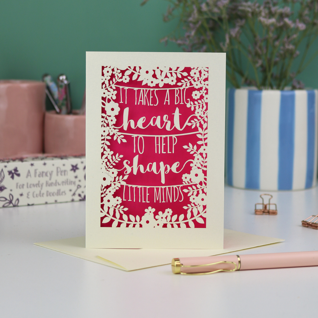 A papercut thank you teacher card with decorative text, 'It takes a big heart to help shape little minds' and floral patterns on a desk with stationery items.