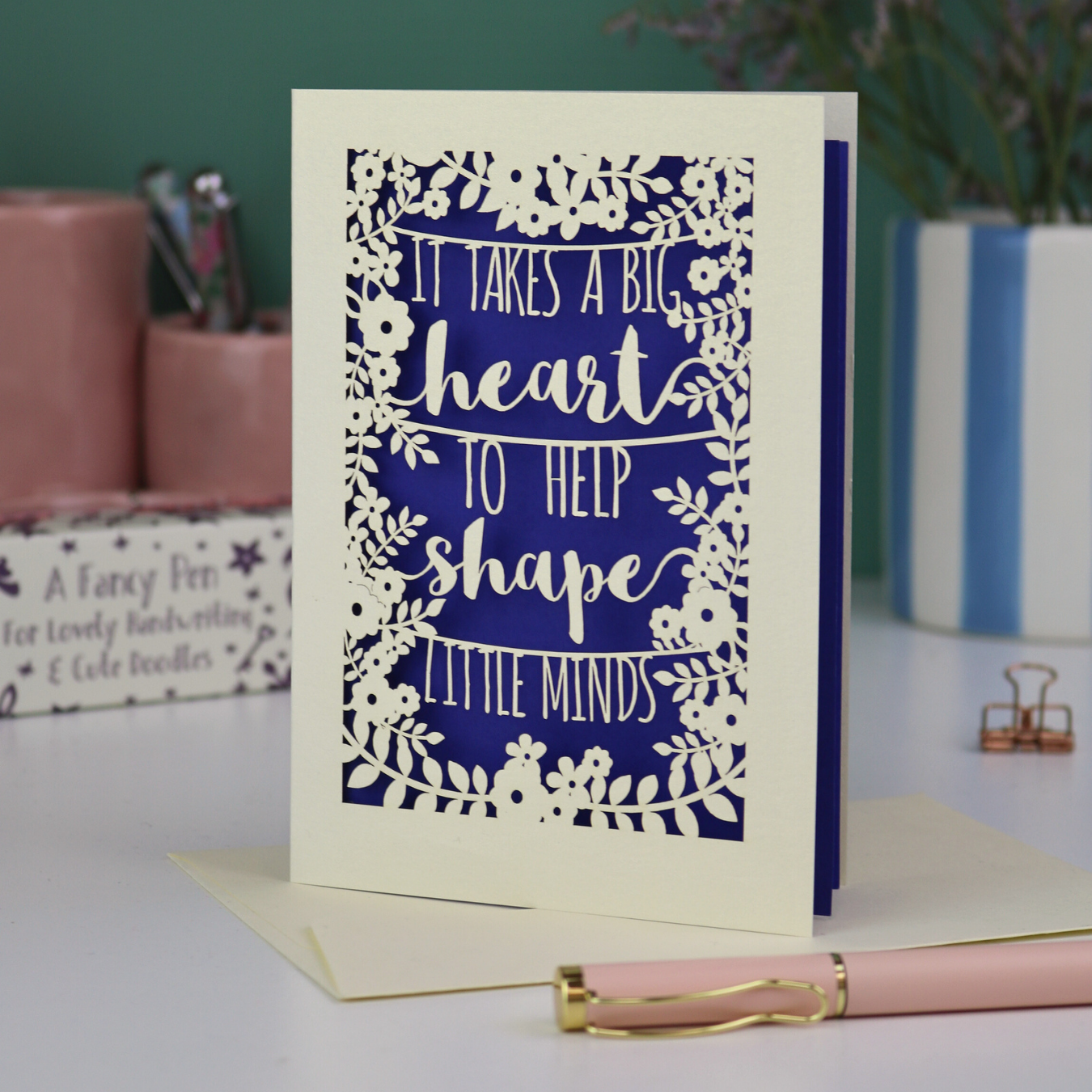 A cut out thank you teacher card with decorative text, 'It takes a big heart to help shape little minds' and floral patterns on a desk with stationery items.