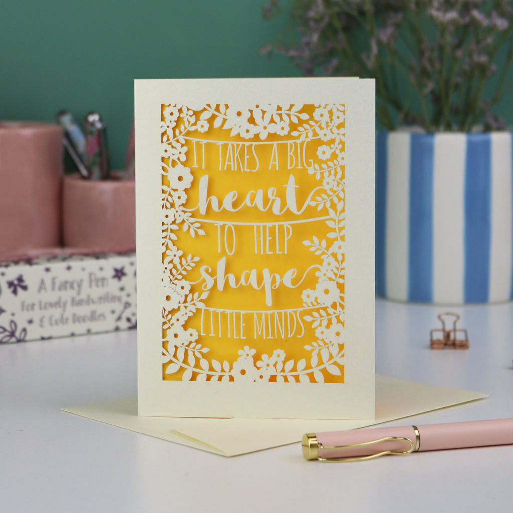 A thoughtful Laser cut thank you teacher card with decorative text, 'It takes a big heart to help shape little minds' and floral patterns on a desk with stationery items.