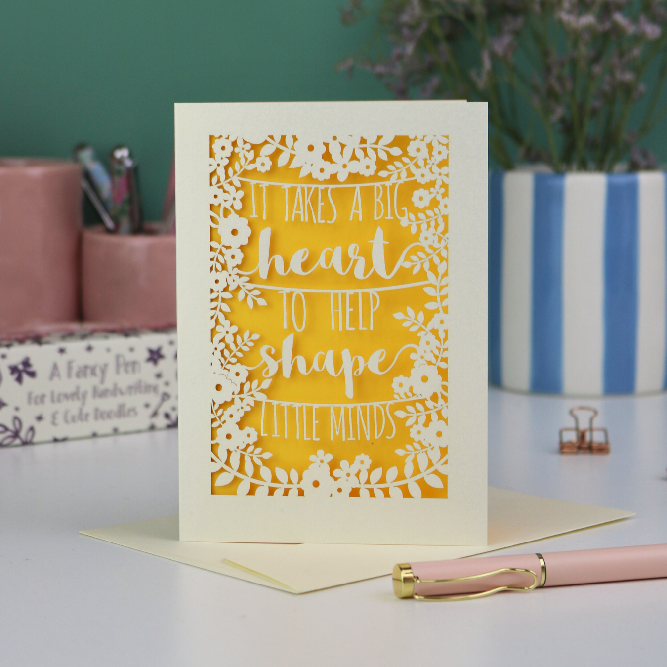 A thoughtful Laser cut thank you teacher card with decorative text, 'It takes a big heart to help shape little minds' and floral patterns on a desk with stationery items.
