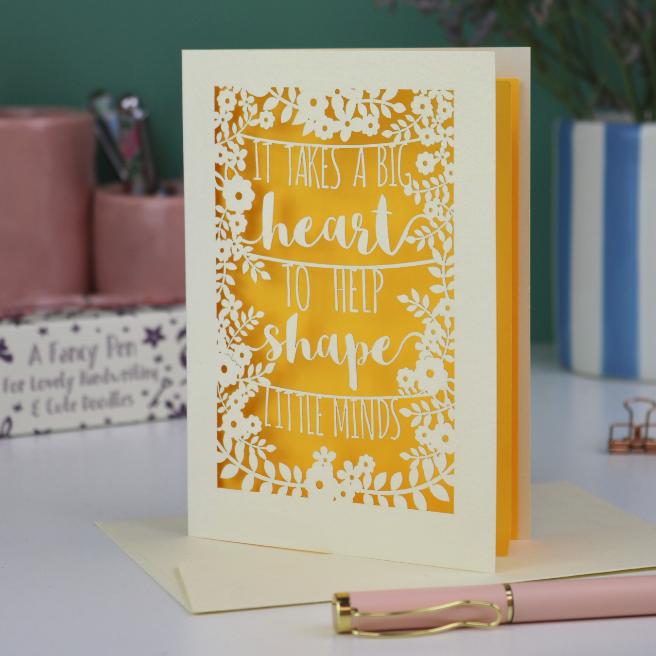 A Laser cut thank you teacher card with decorative text, 'It takes a big heart to help shape little minds' and floral patterns on a desk with stationery items.