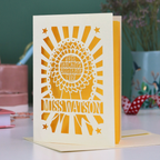 A laser cut thank you teaching assistant card with cut out rosette design and text "Best Teaching Assistant Ever" and personalised with the name "Miss Watson", on a desk with stationery items