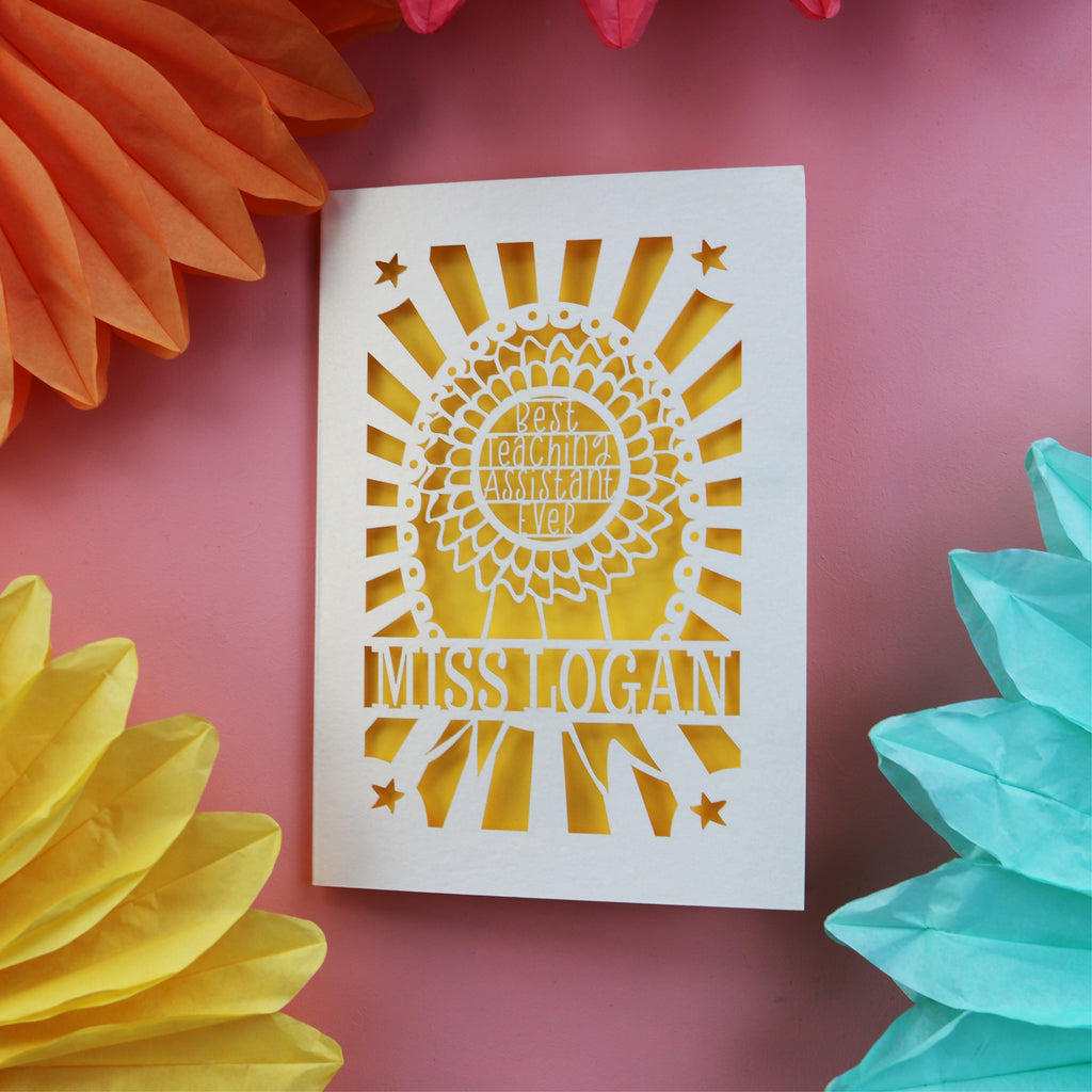 A cut out thank you teacher card for a teaching assistant. Card has a rosette with "Best teaching assistant ever" and is personalised with their name