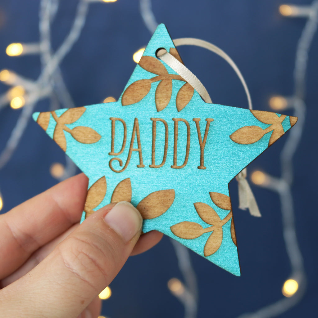 A hand holding a teal star shaped Christmas decoration, personalised with a name.