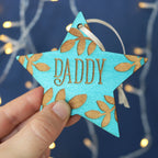 A hand holding a teal star shaped Christmas decoration, personalised with a name.