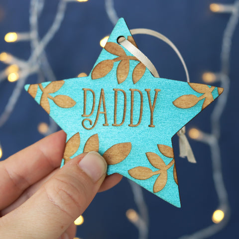 A hand holding a teal star shaped Christmas decoration, personalised with a name. - 