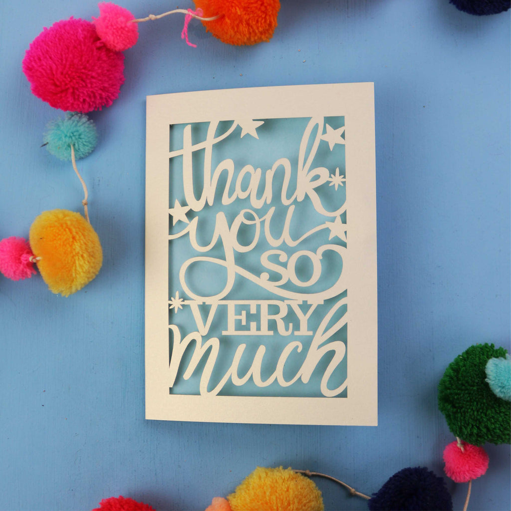 Laser cut thanks card with 'Thank you so very much' text on a blue background with colourful pom-poms.