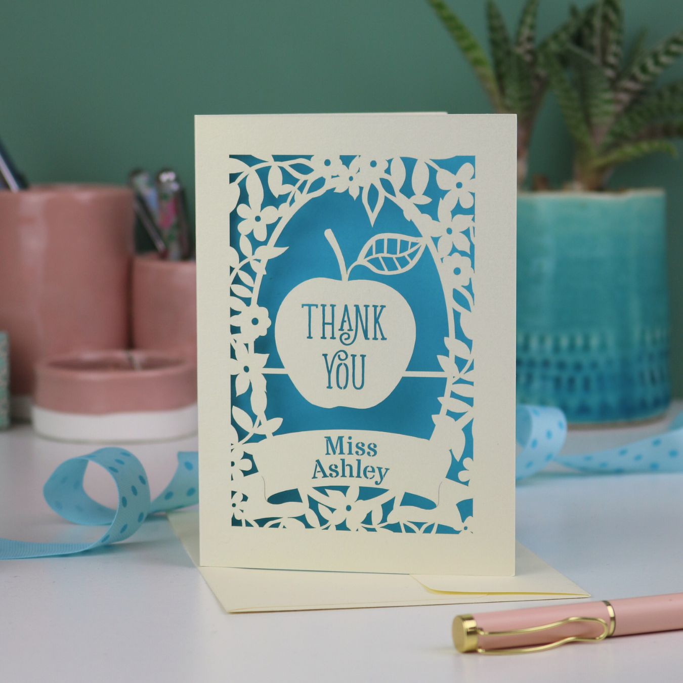 Laser cut Thank you teacher card with apple design, decorative border and text, 'Thank You Miss Ashley',  on a desk with stationery items.