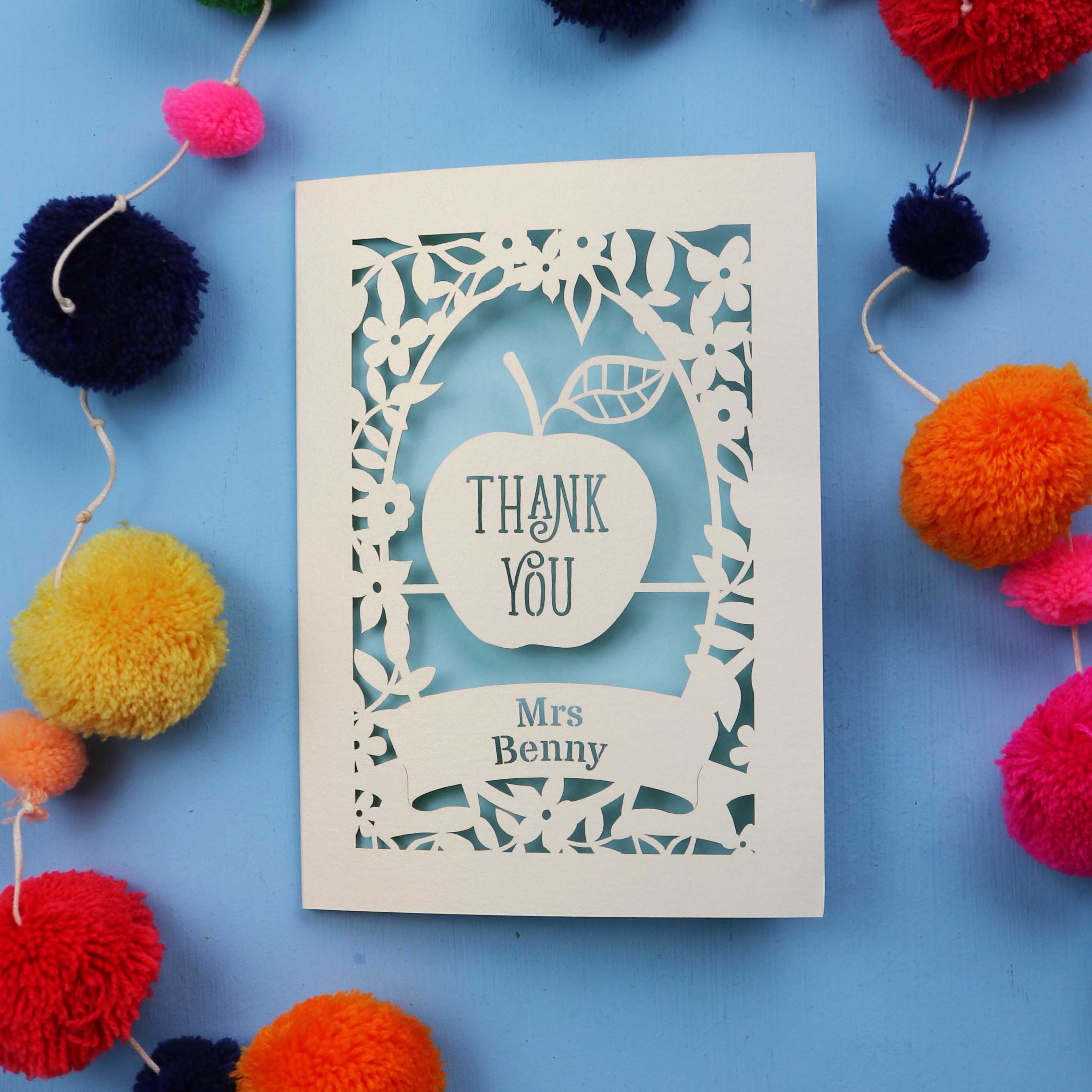 Personalised teacher card with 'Thank You Mrs Benny' text surrounded by colourful pom-poms on a blue background