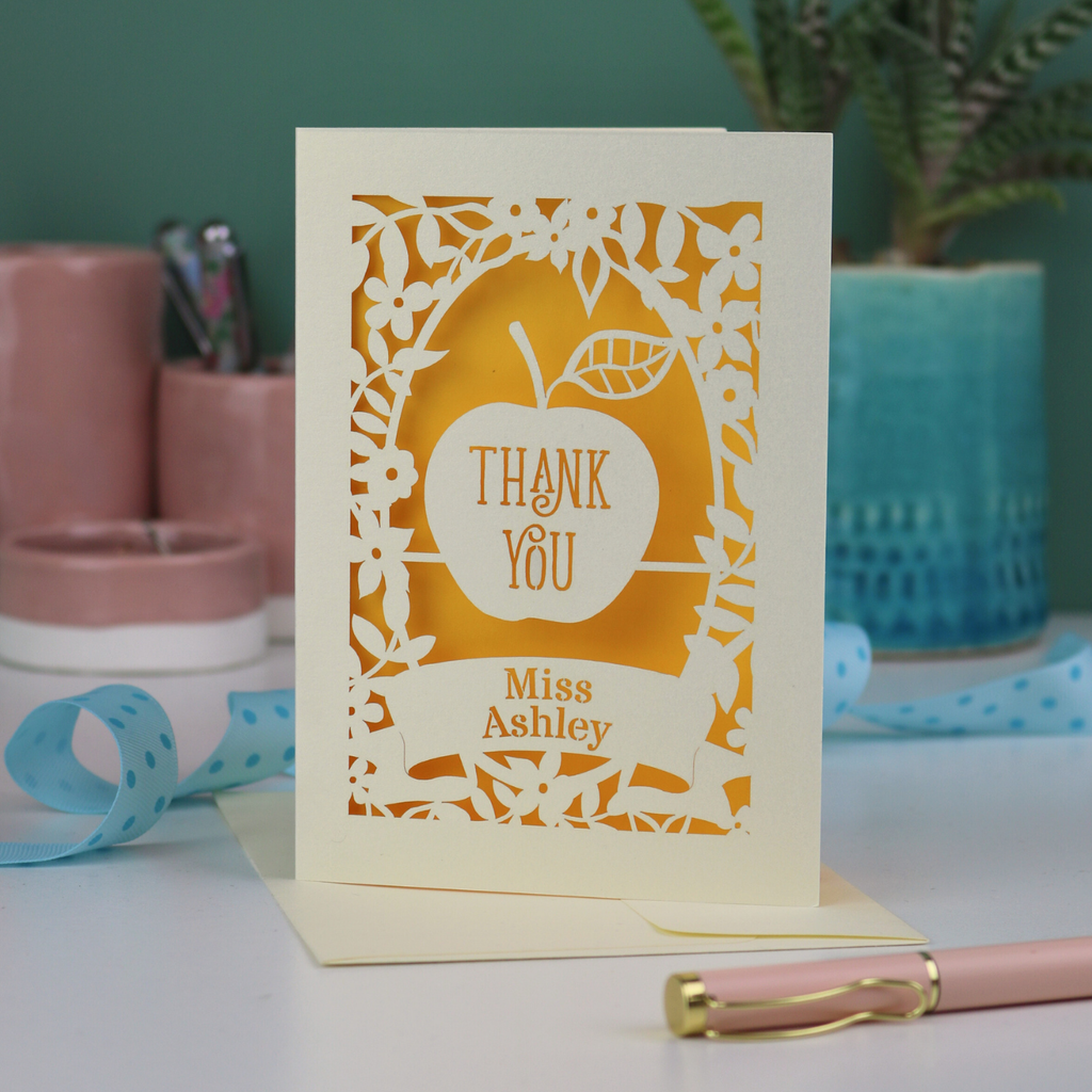 Unique Thank you teacher card with apple design, decorative border and text, 'Thank You Miss Ashley',  on a desk with stationery items.
