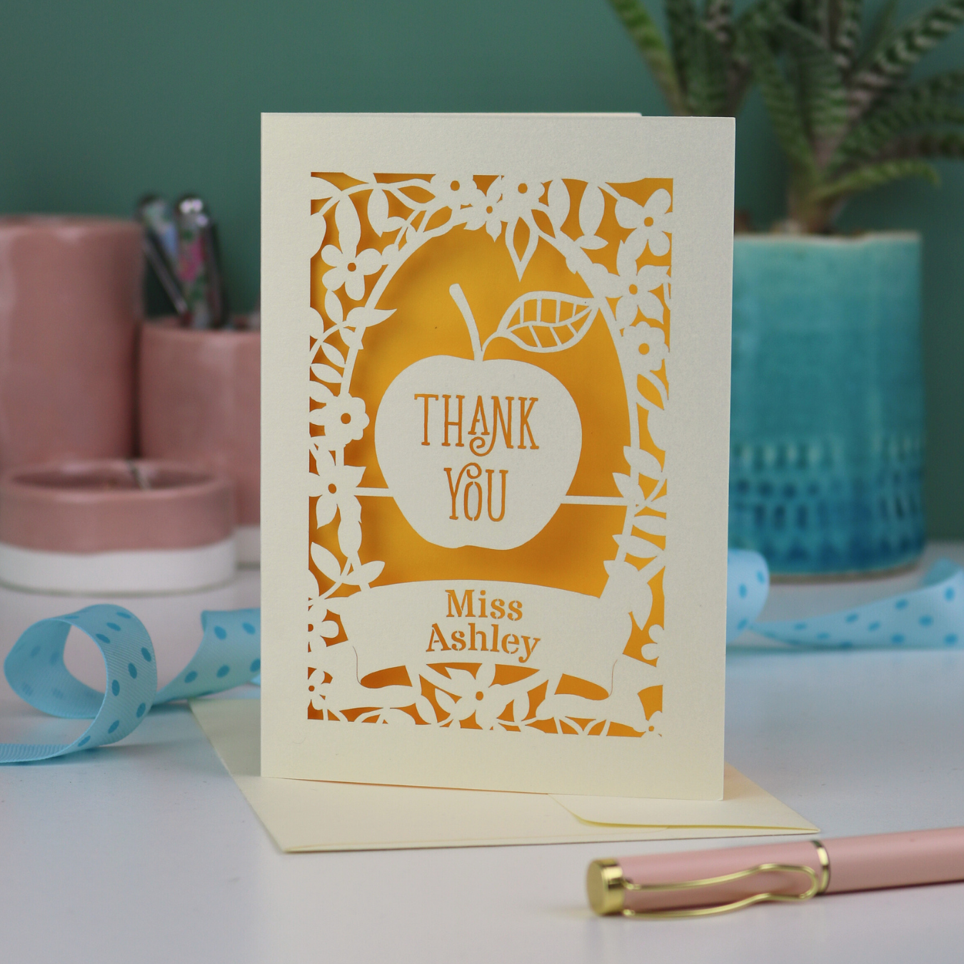 Unique Thank you teacher card with apple design, decorative border and text, 'Thank You Miss Ashley',  on a desk with stationery items.
