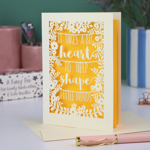A papercut thank you teacher card with decorative text, 'It takes a big heart to help shape little minds' and floral patterns on a desk with stationery items.