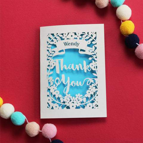 Personalised papercut thank you card. Cut from cream card with a peacock blue insert paper. Shows the words Thank You surrounded with flowers and a banner to personalise with a name.