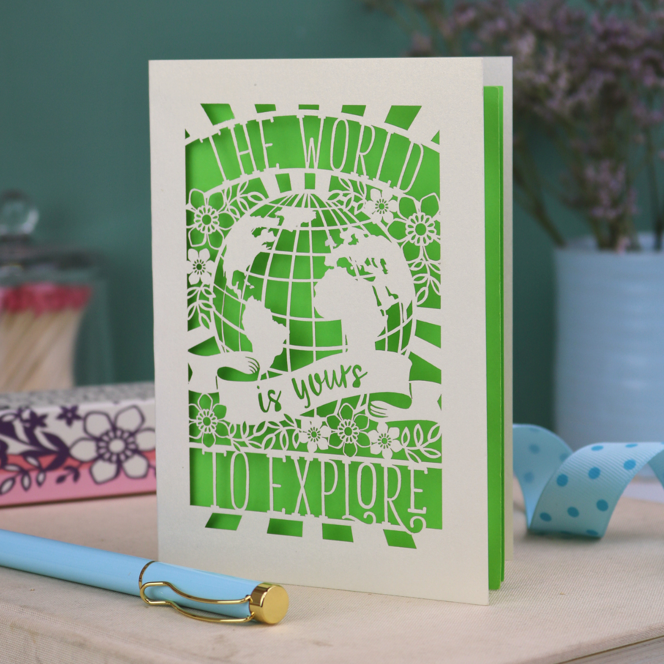 Card with laser-cut design and 'The world is yours to explore' text, accompanied by a pen and stationery items on a desk.