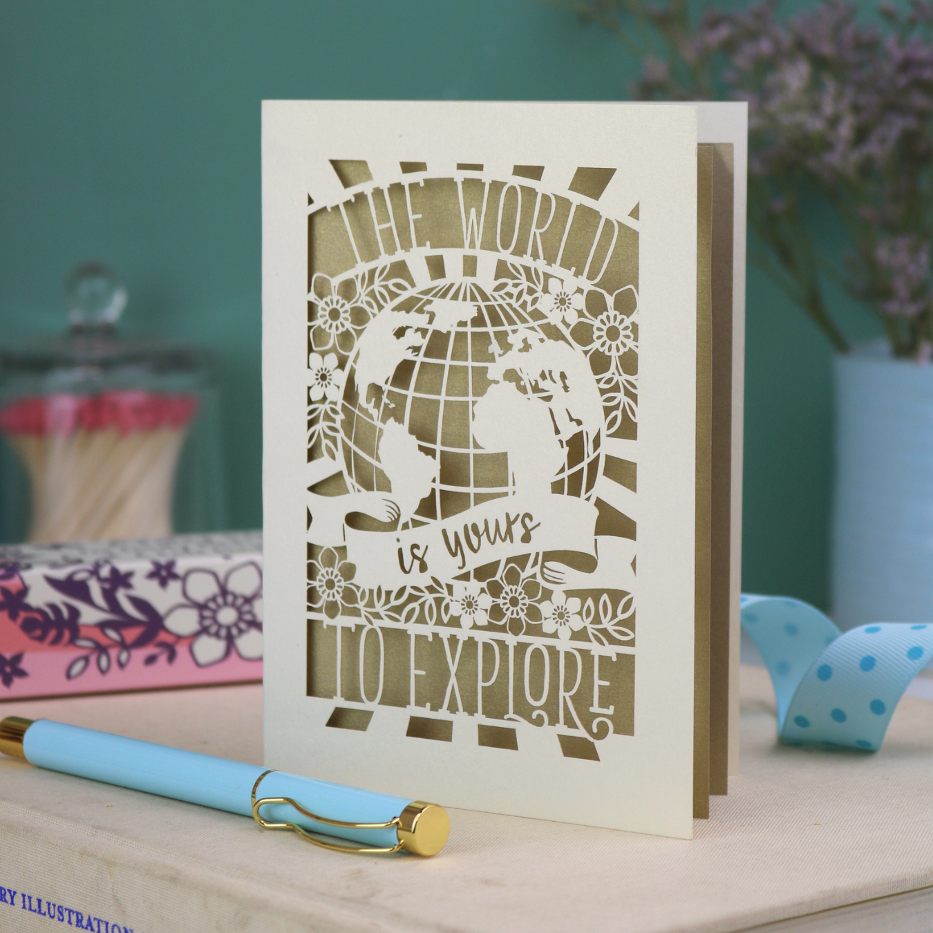 Laser cut greetings Card with globe design and 'The world is yours to explore' text, accompanied by a pen and stationery items on a desk.