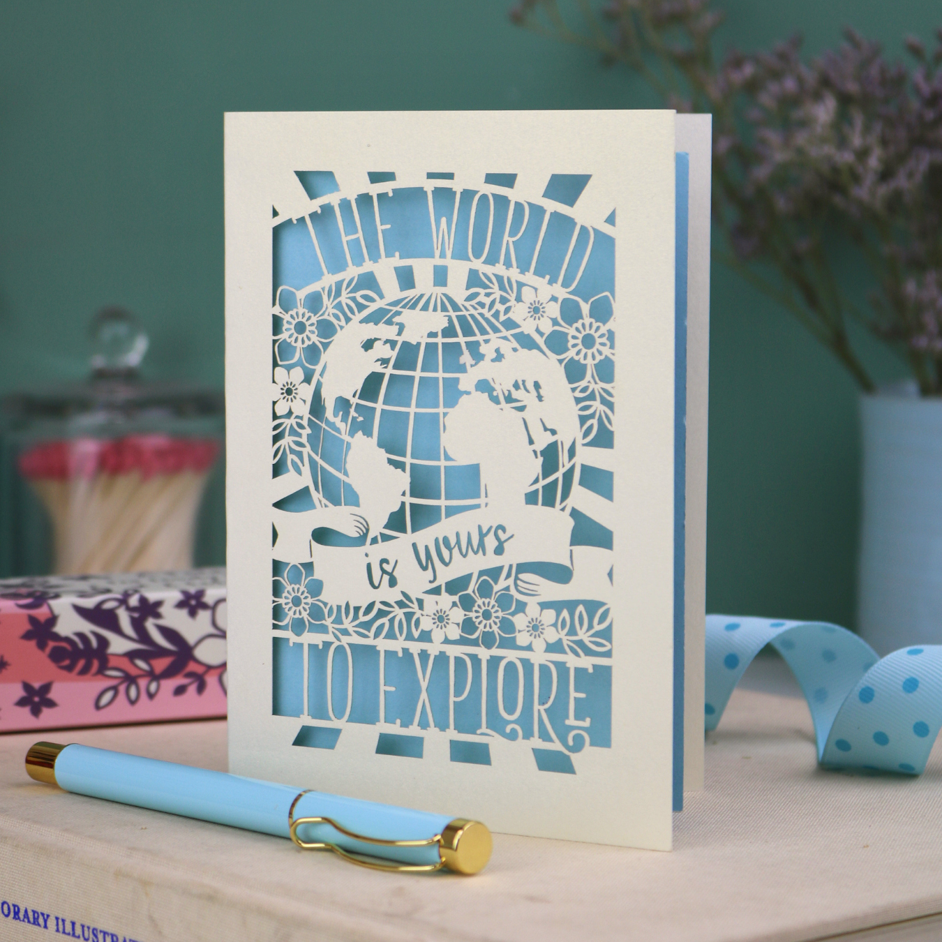 Laser cut greeting card with globe design and 'The world is yours to explore' text, accompanied by a pen and stationery items on a desk.