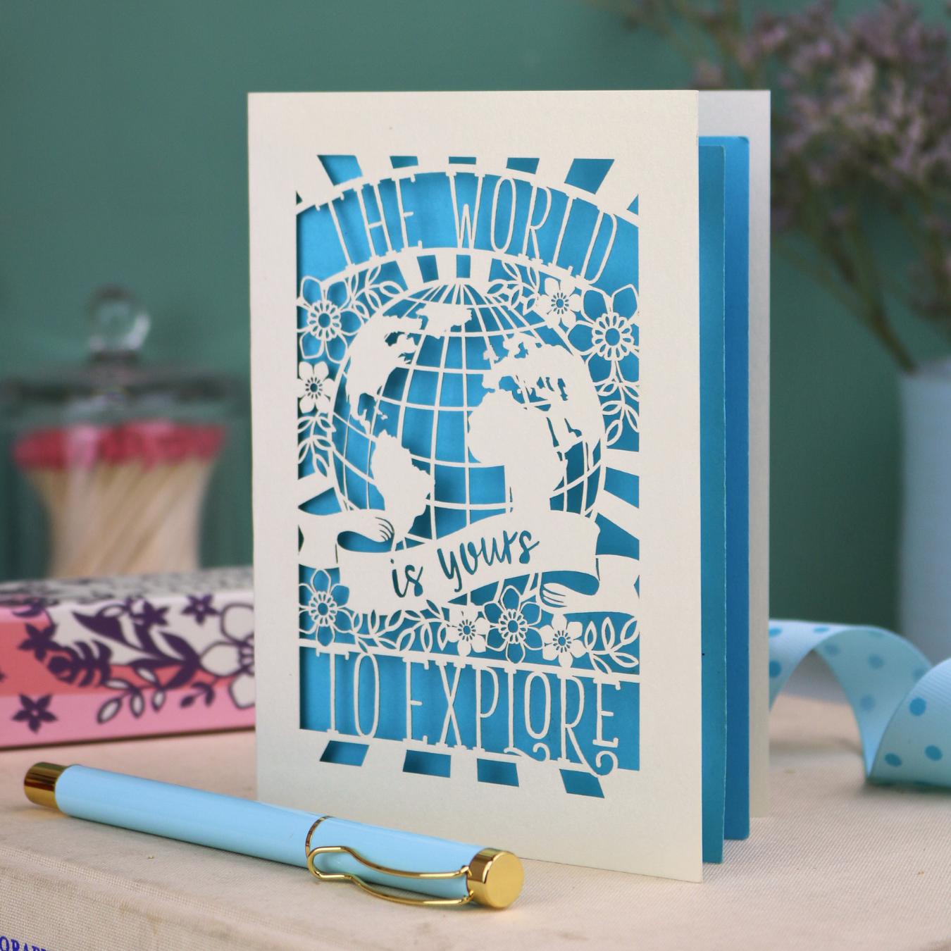Card with laser-cut design and 'The world is yours to explore' text, accompanied by a pen and stationery items on a desk.