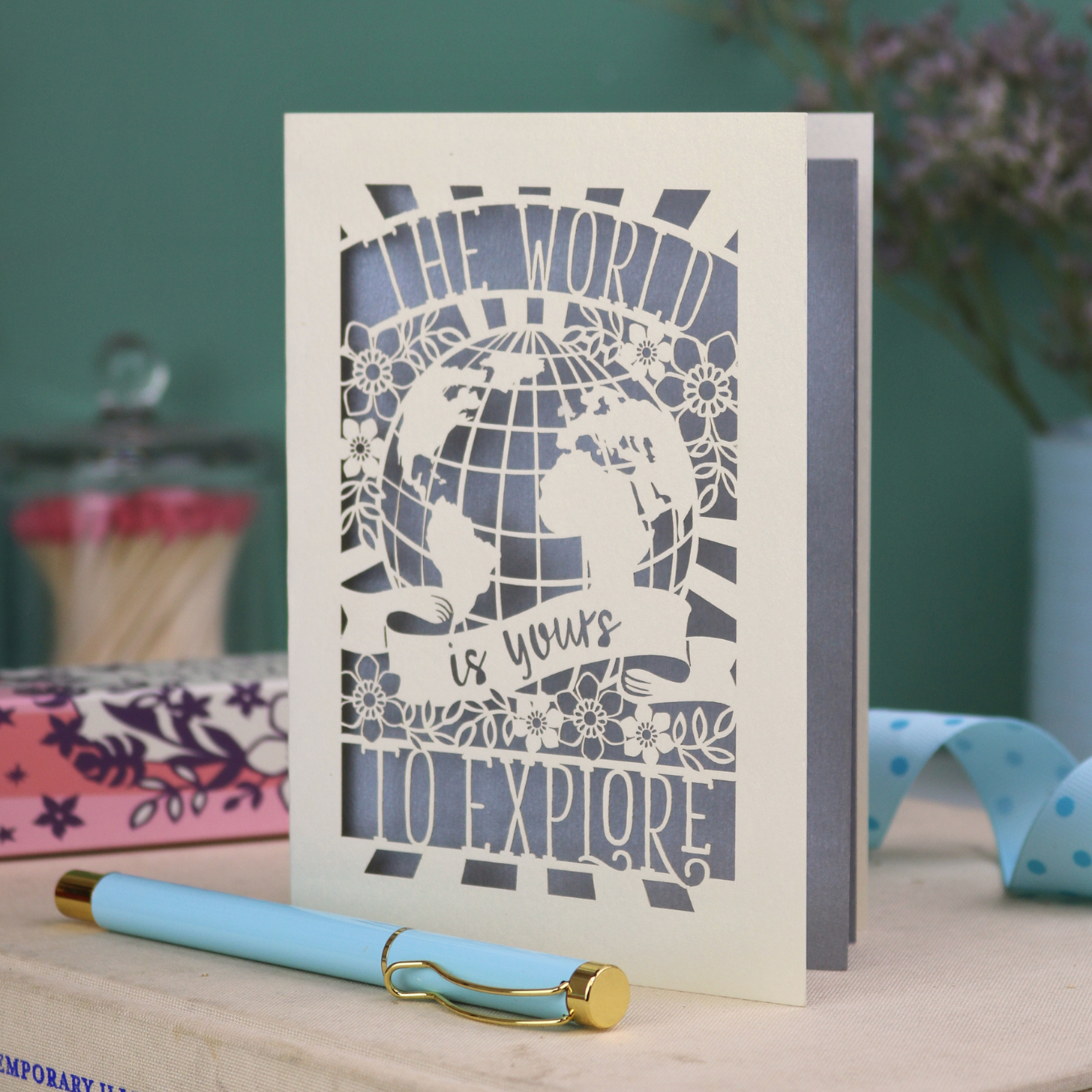 Laser cut greetings Card with globe design and 'The world is yours to explore' text, accompanied by a pen and stationery items on a desk.