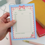 A hand holding a light blue daily planner notepad with a pink wavy border and floral accents. The pad is labeled 'Today's Plan' with sections for date, schedule, to-do list, and reminders.