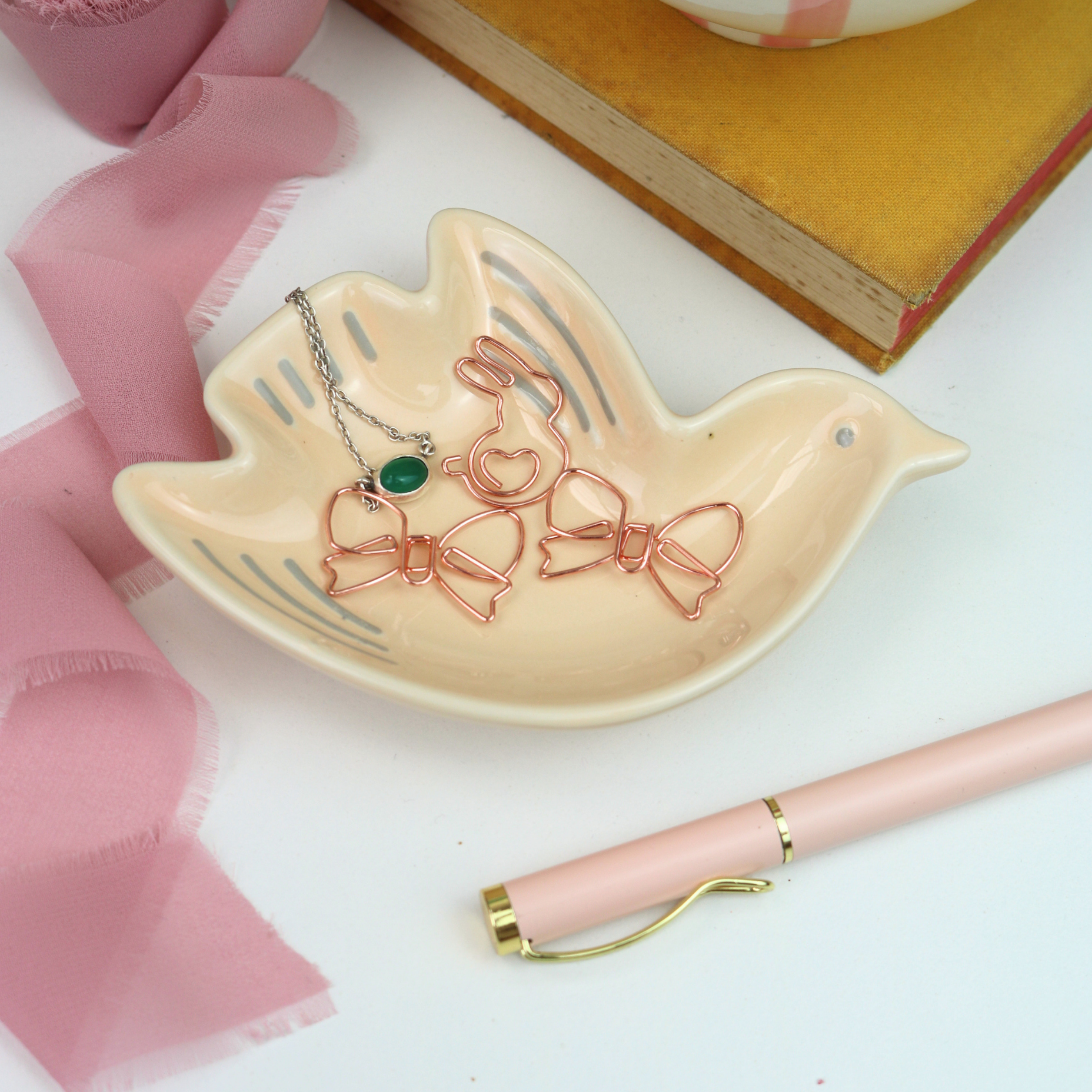 Decorative dish shaped like a bird with jewelry and hair clips, pink pen and fabric in the background.