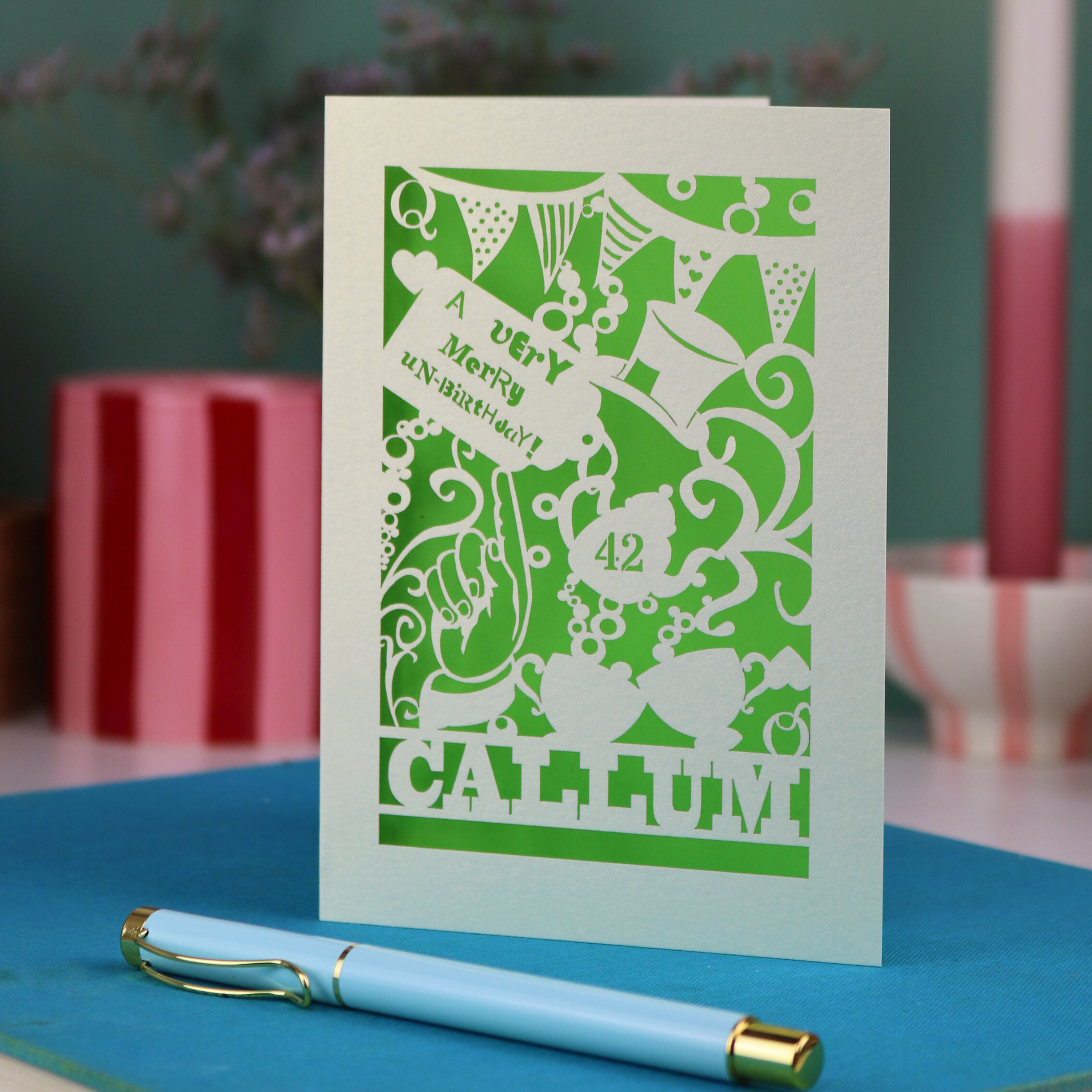 A laser cut 'Merry Un-Birthday' card with pen and stationery items on a blue surface.