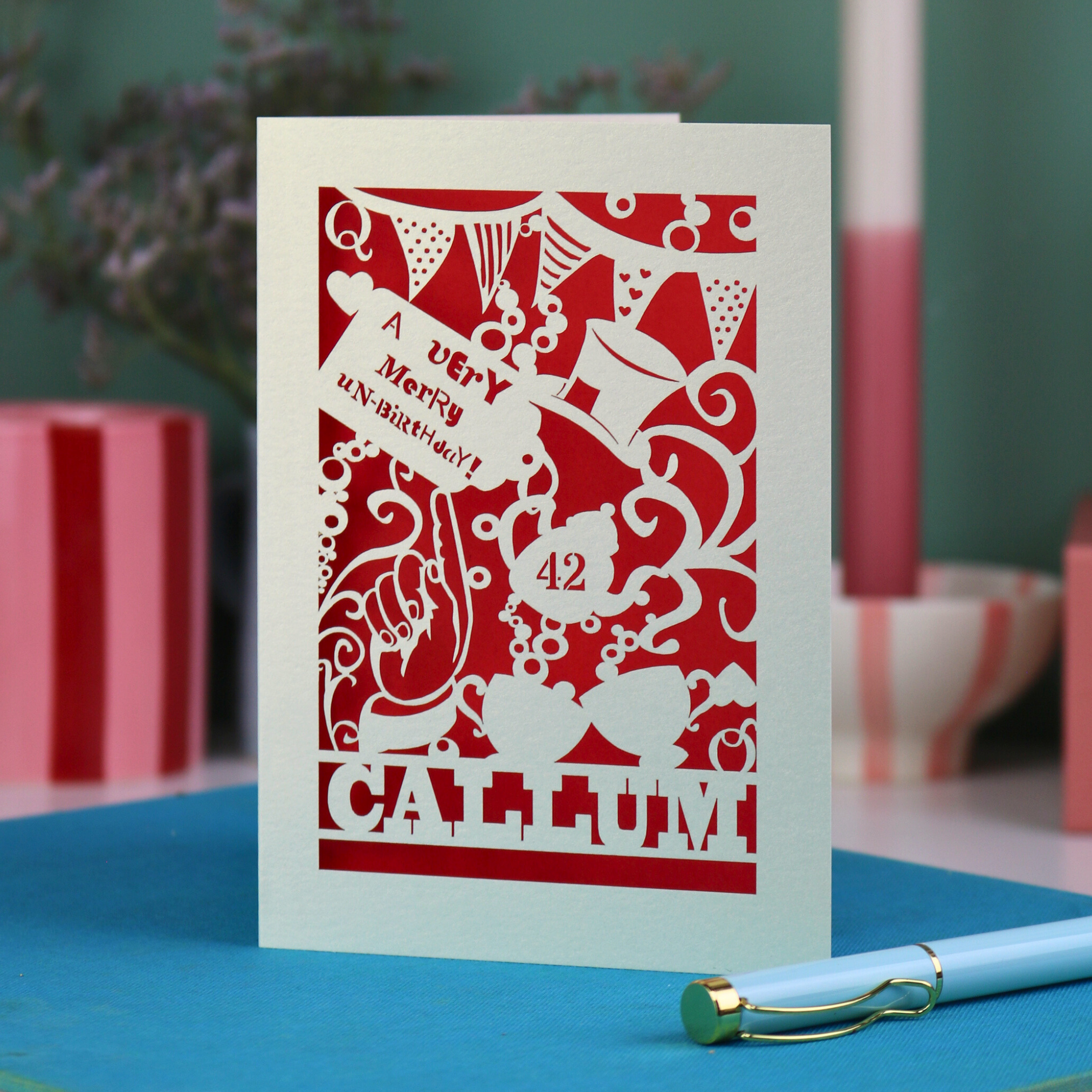 A laser cut 'Merry Un-Birthday' card with pen and stationery items on a blue surface.