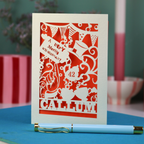 A laser cut 'Merry Un-Birthday' card with pen and stationery items on a blue surface.