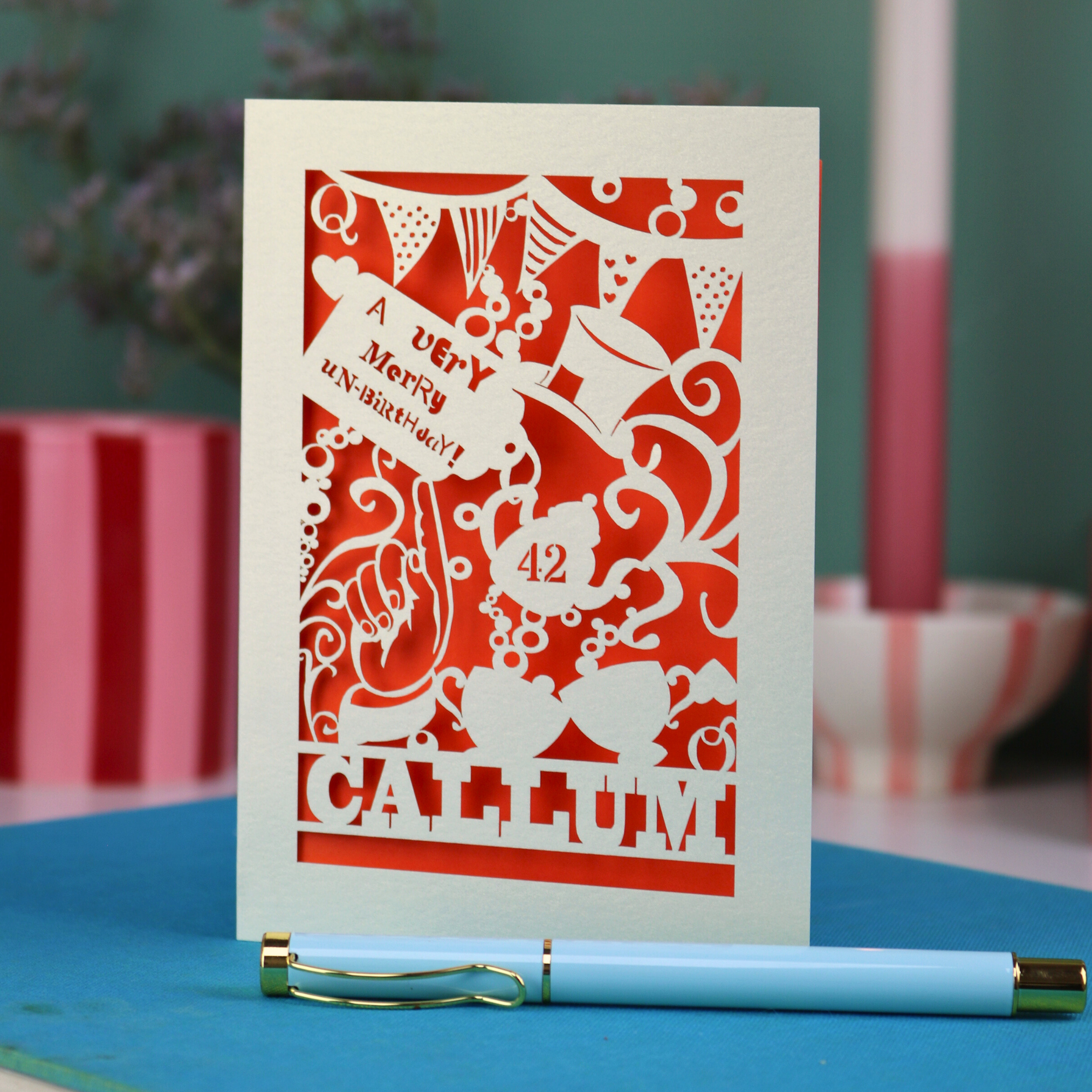A laser cut 'Merry Un-Birthday' card with pen and stationery items on a blue surface.