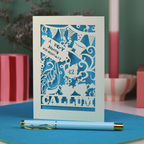 A laser cut 'Merry Un-Birthday' card with pen and stationery items on a blue surface.