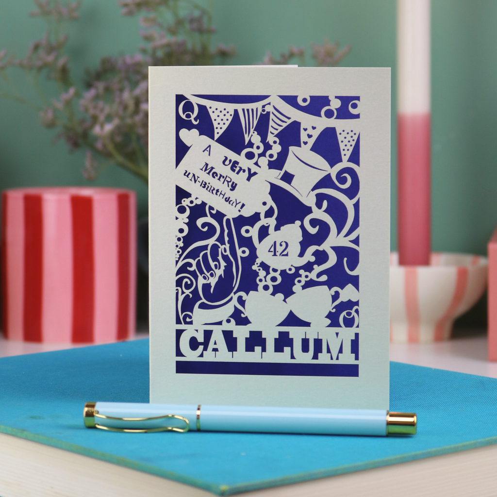 A laser cut 'Merry Un-Birthday' card with pen and stationery items on a blue surface.