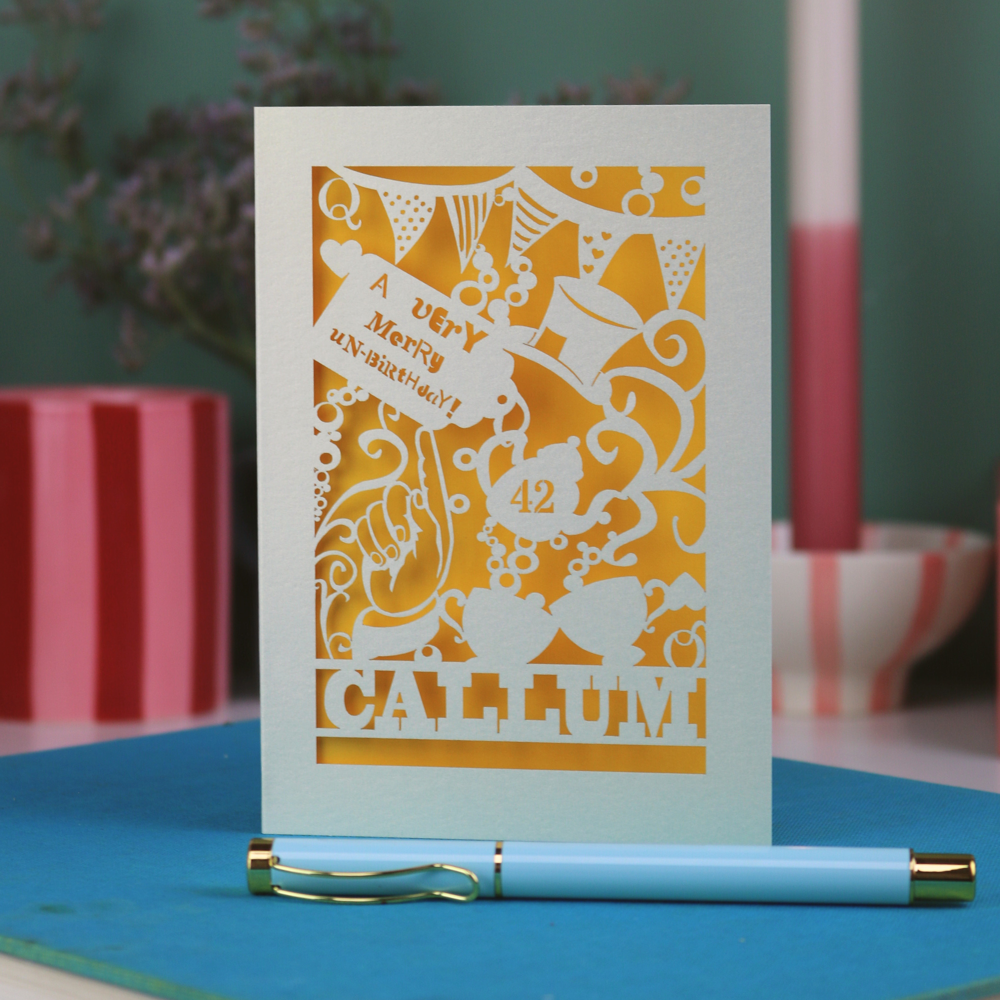 A laser cut 'Merry Un-Birthday' card with pen and stationery items on a blue surface.