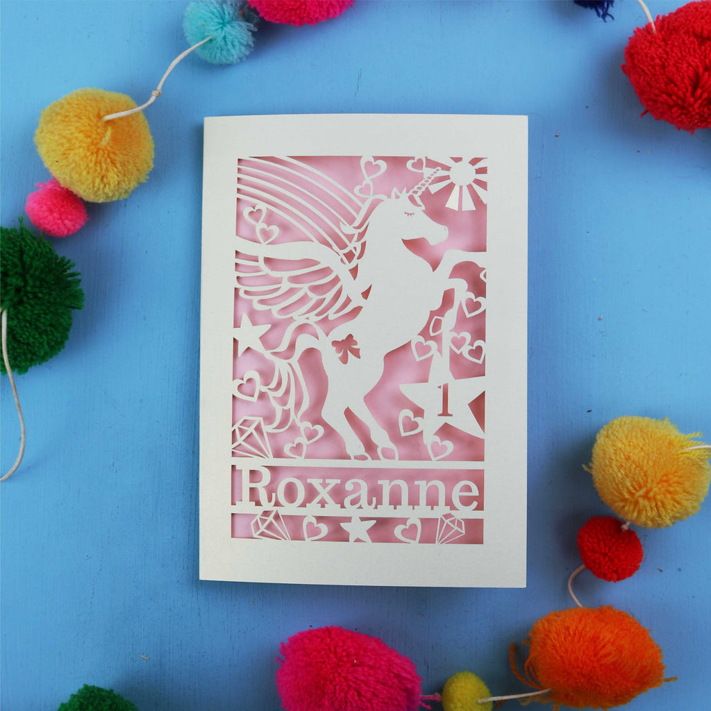 Paper cut out 1st birthday card with a unicorn with 'Roxanne' on a blue background with colourful pom-poms.