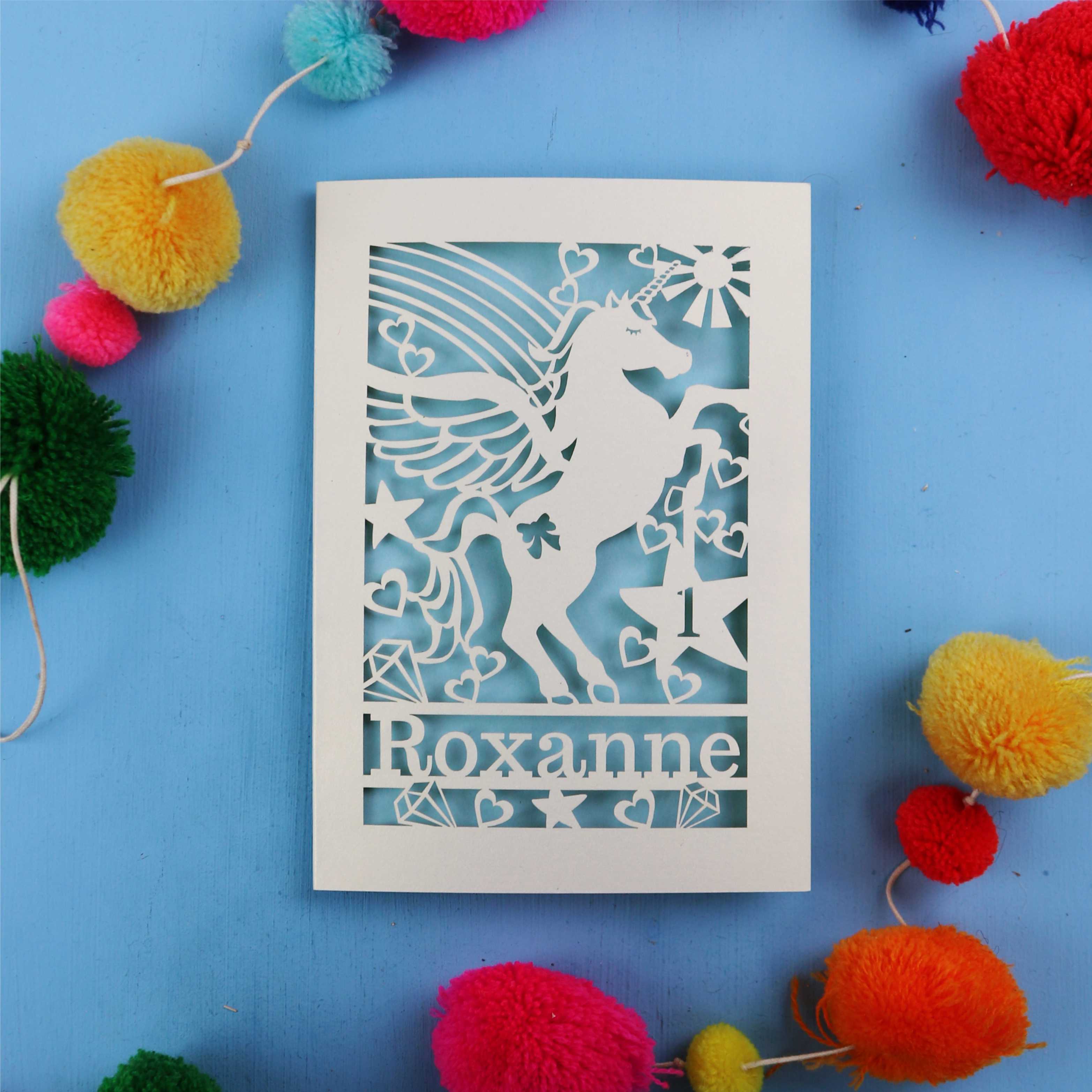 Paper cut out 1st birthday card with a unicorn with 'Roxanne' on a blue background with colourful pom-poms.