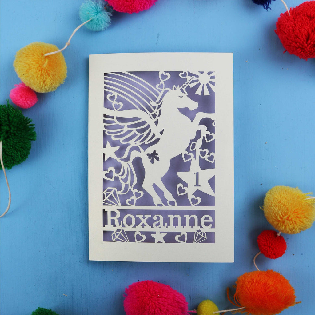 Paper cut out 1st birthday card with a unicorn with 'Roxanne' on a blue background with colourful pom-poms.