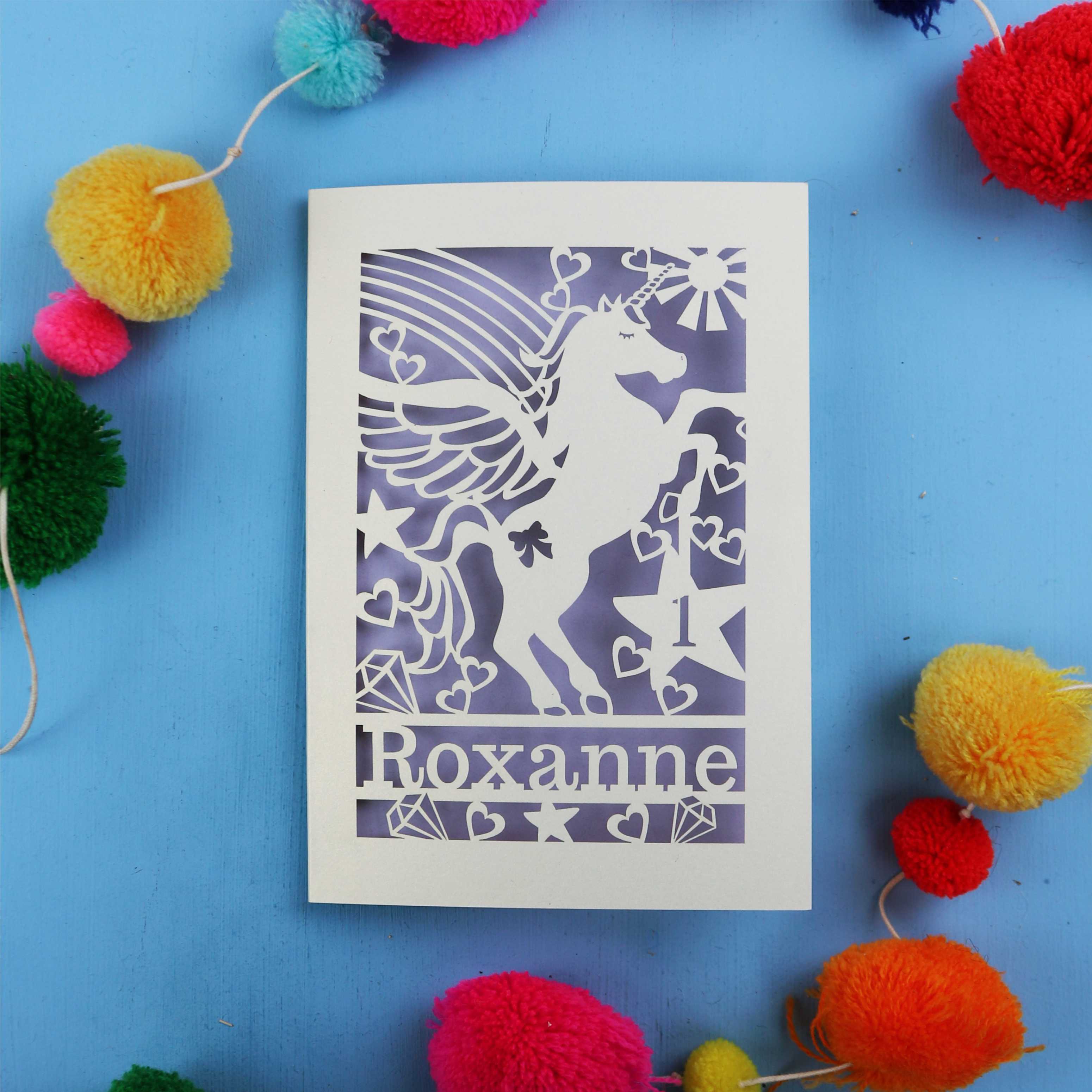 Paper cut out 1st birthday card with a unicorn with 'Roxanne' on a blue background with colourful pom-poms.