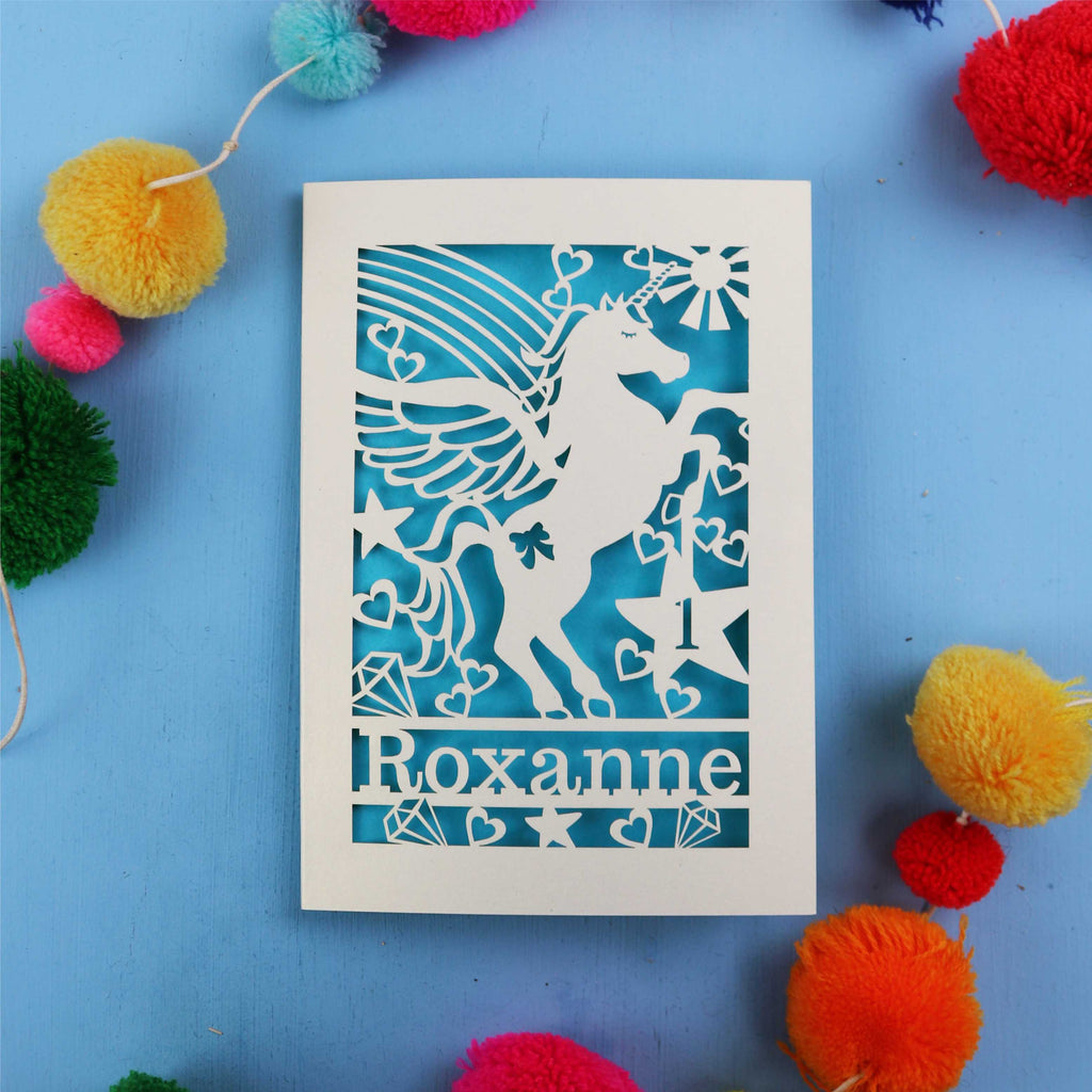 Paper cut out 1st birthday card with a unicorn with 'Roxanne' on a blue background with colourful pom-poms.