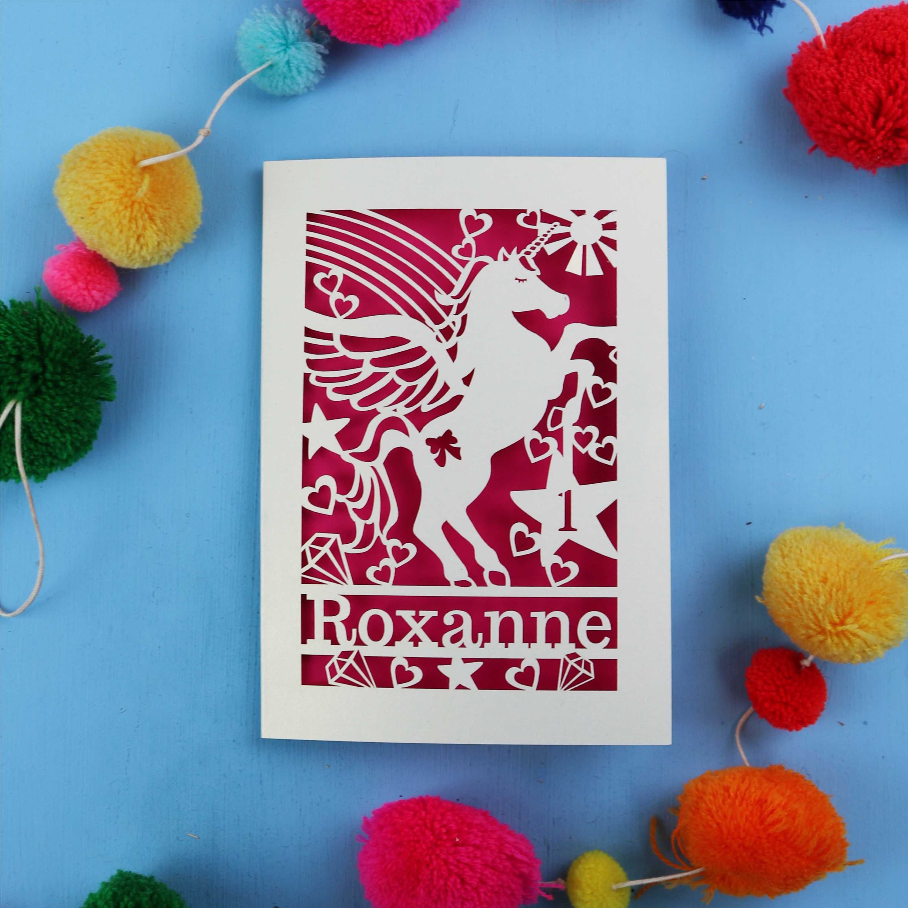 1st Birthday Card with unicorn design and 'Roxanne' text on a blue background with colourful pom-poms.