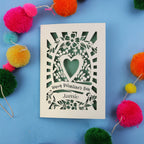 Decorative laser cut Valentine's card with floral heart design on a blue background with colourful pom-poms.
