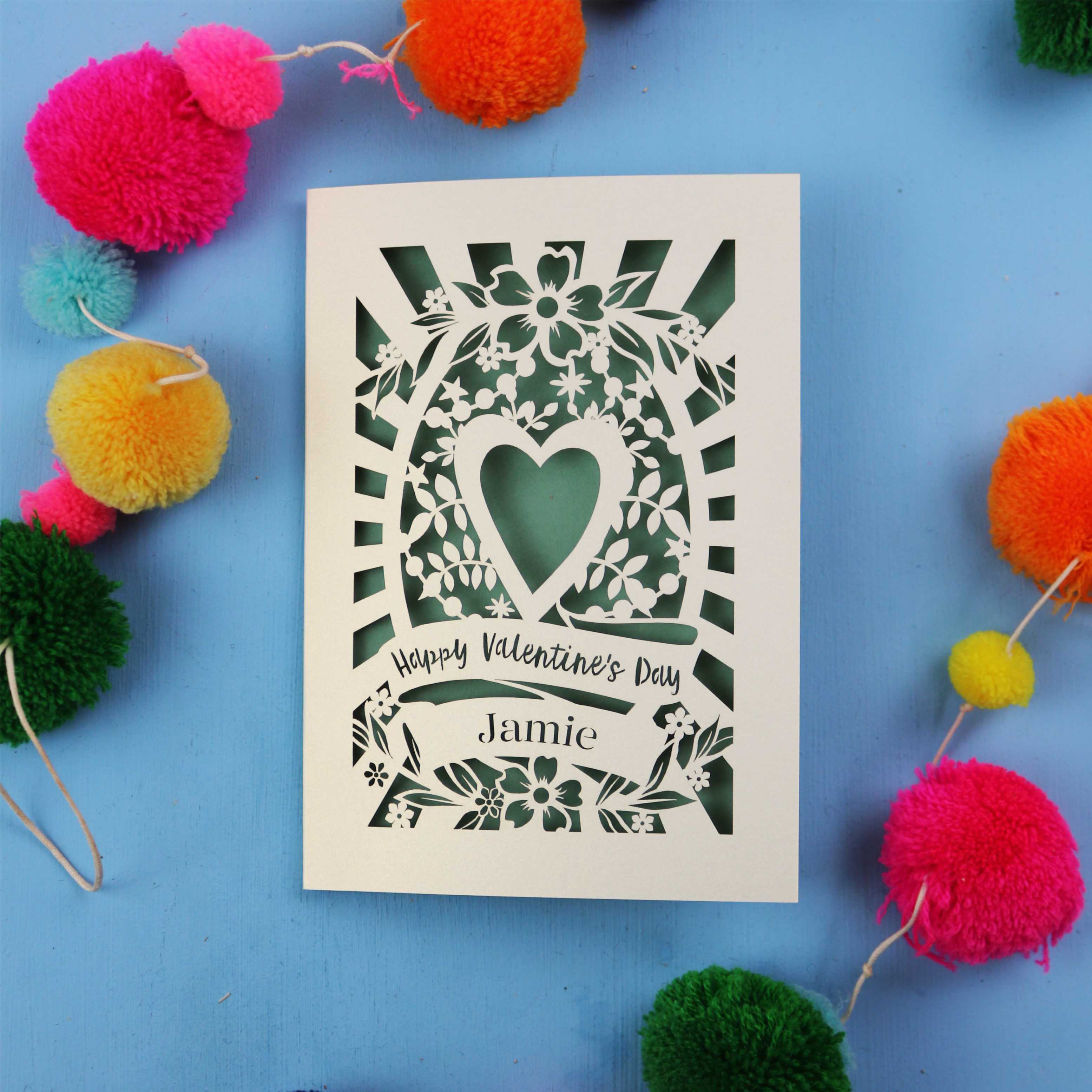 Decorative laser cut Valentine's card with floral heart design on a blue background with colourful pom-poms.