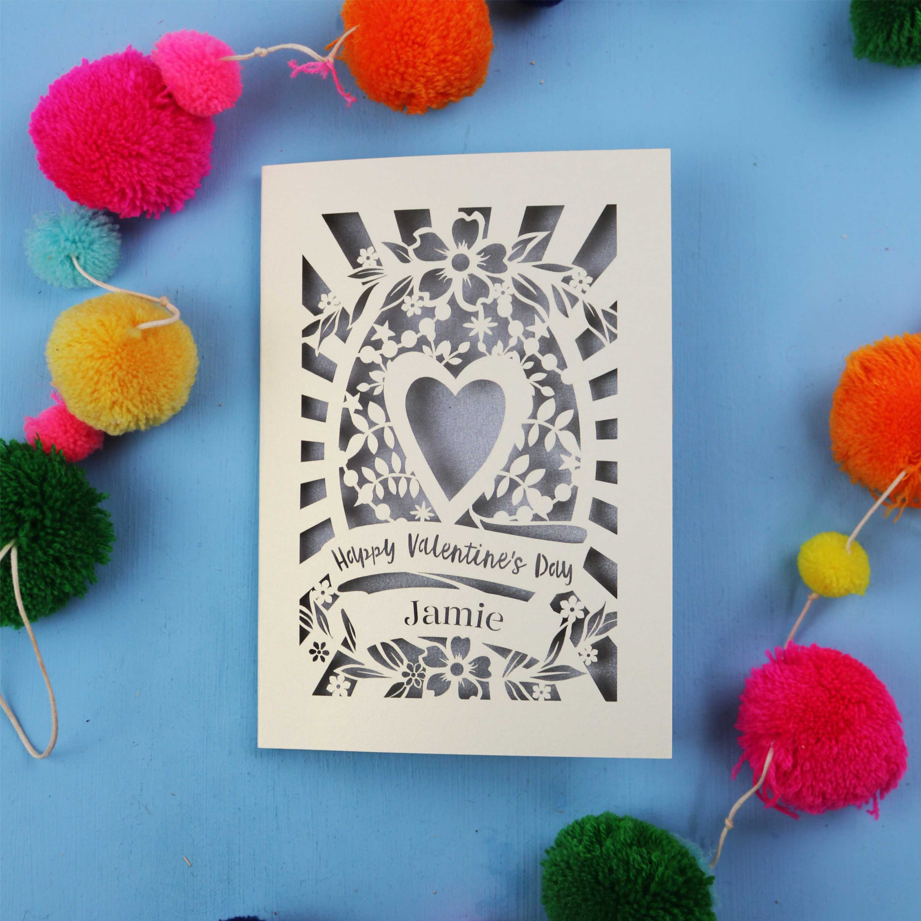 Decorative Valentine's card with floral heart design on a blue background with colourful pom-poms.