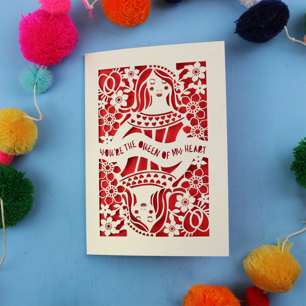 Valentine's Card with laser cut design of the queen of hearts on a blue background with colourful pom-poms.
