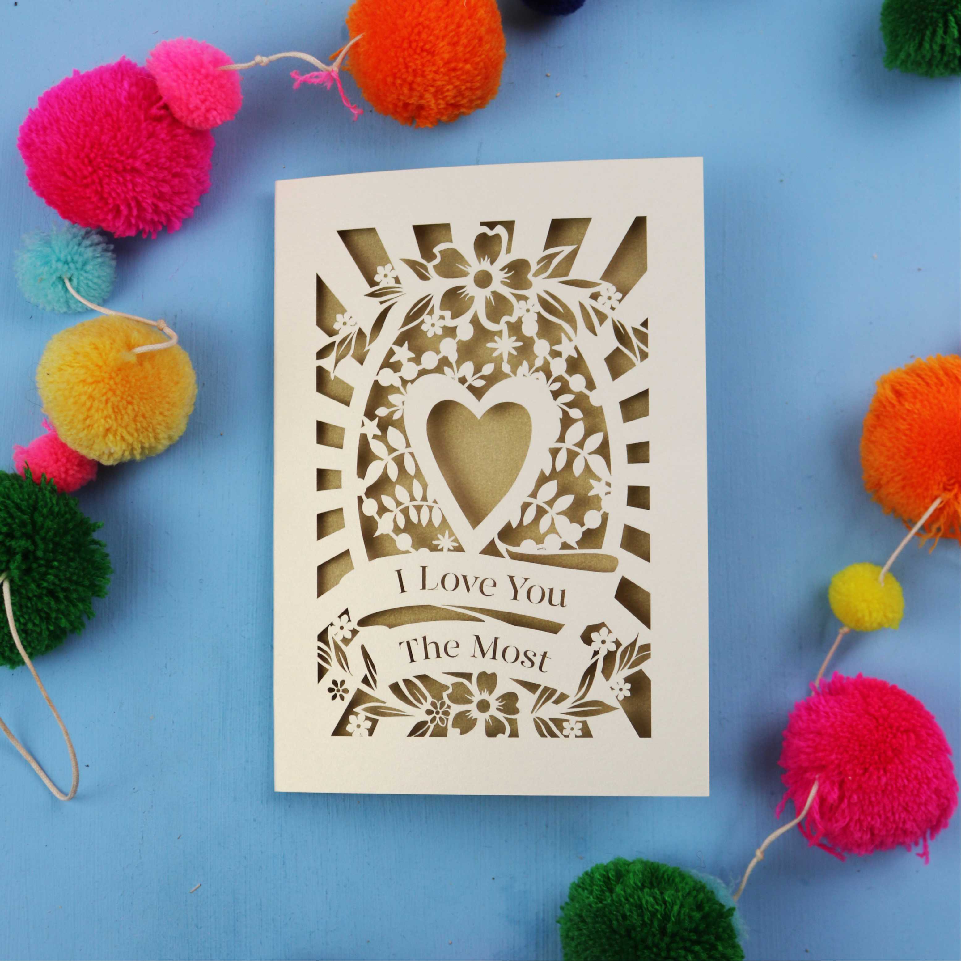 Laser cut Valentines Card with 'I Love You The Most' design on a blue background with colourful pom-poms.