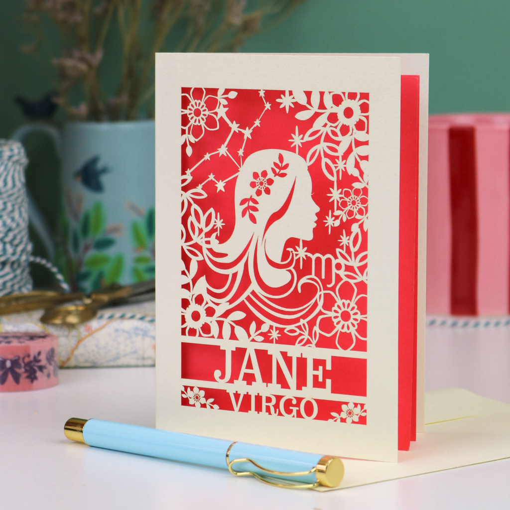 A personalised laser-cut Virgo birthday card card with a silhouette of the Virgo 'maiden', the constellation, zodiac symbol and text 'Virgo', surrounded by floral patterns, displayed on a white surface with a light blue pen in front.