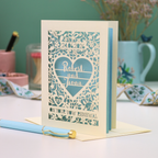 Laser-cut vow renewal card with decorative design and text on a table with stationery items.