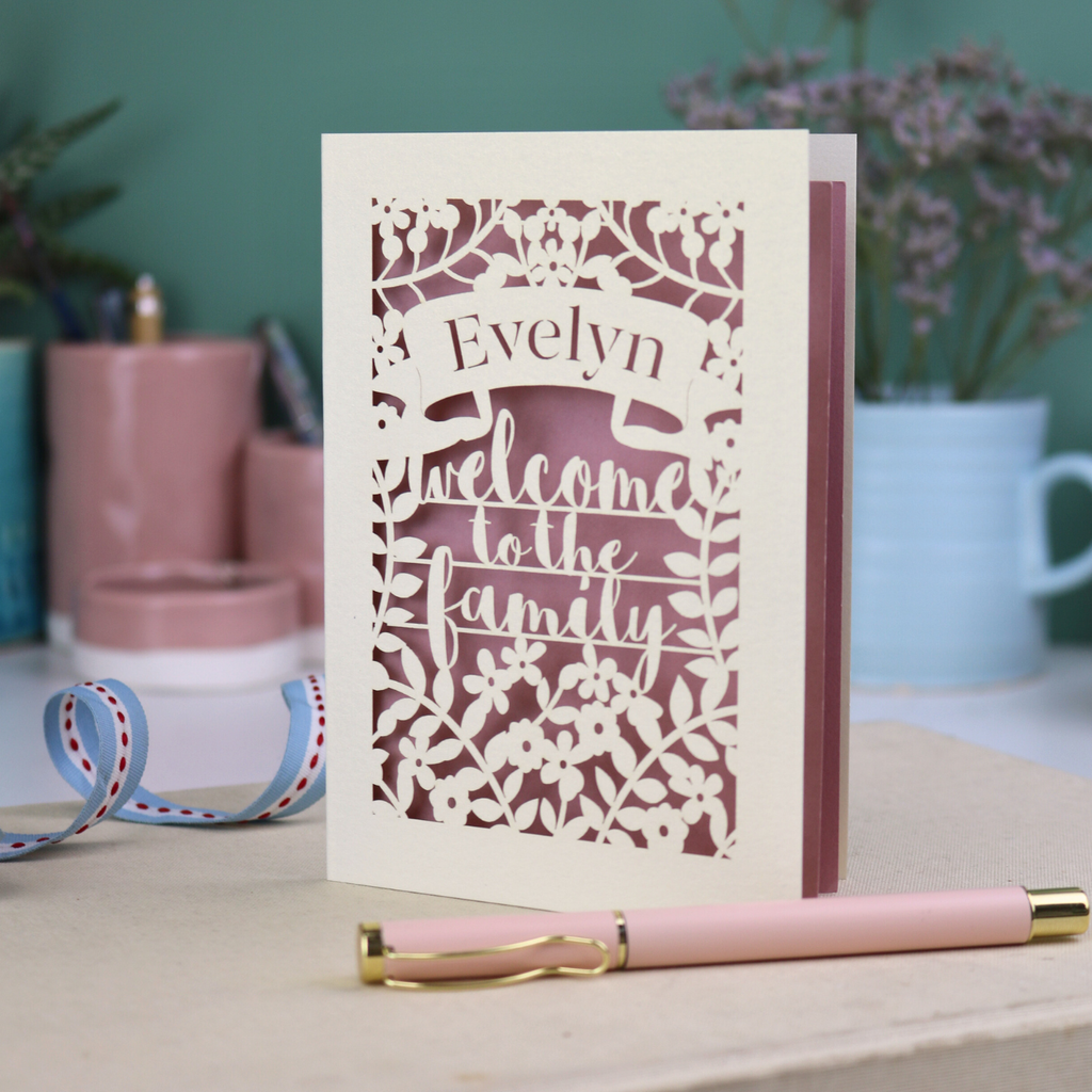 A floral laser cut wedding card personalised with a name and the text "Welcome to the Family", standing on a desk with stationery items.