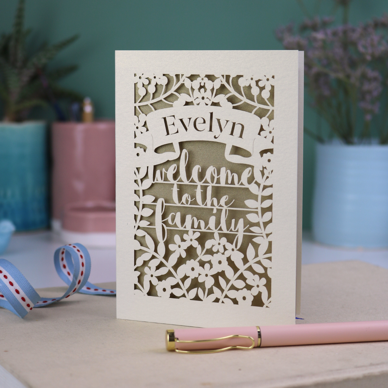 A floral laser cut wedding card personalised with a name and the text "Welcome to the Family", standing on a desk with stationery items.