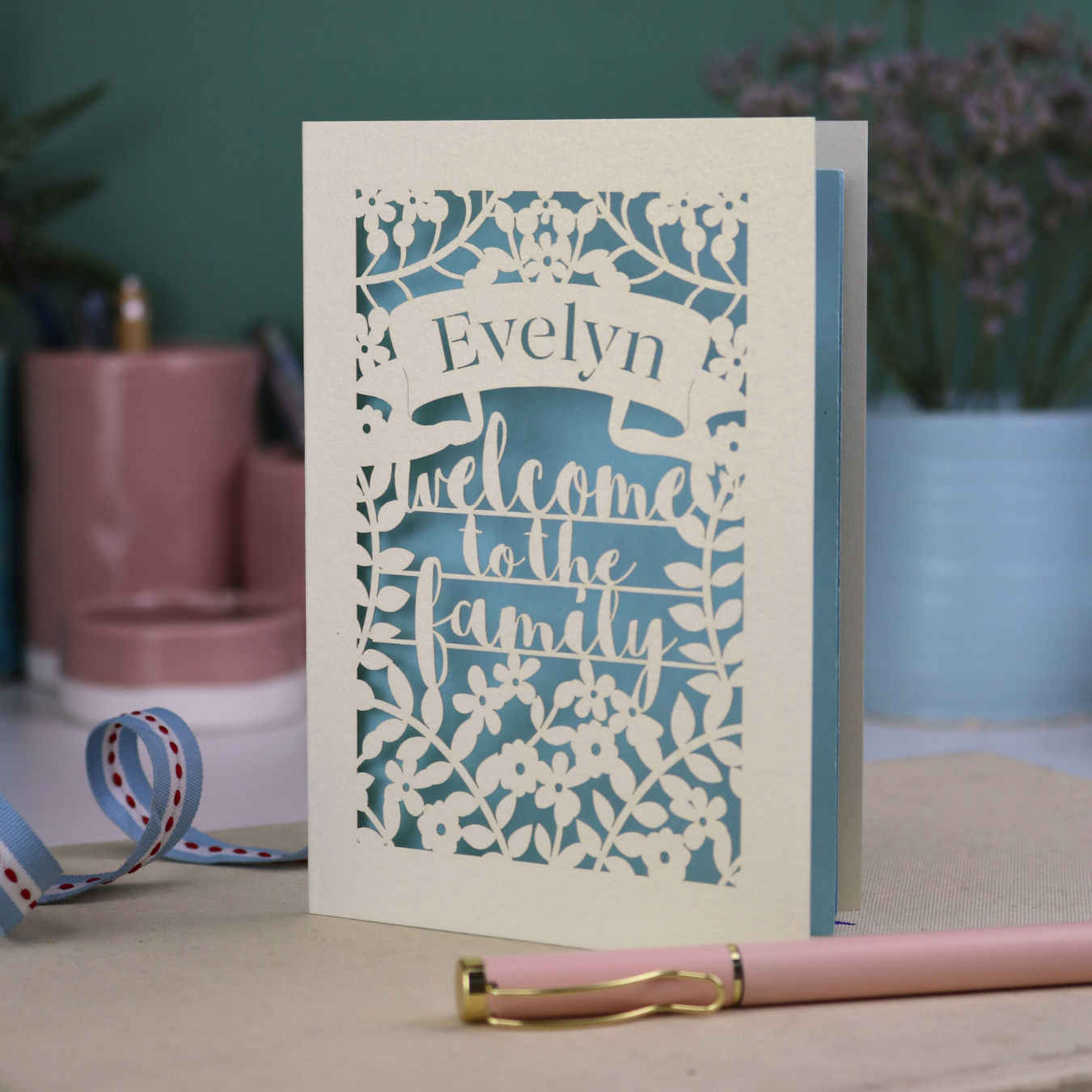 A floral laser cut wedding card personalised with a name and the text "Welcome to the Family", standing on a desk with stationery items.